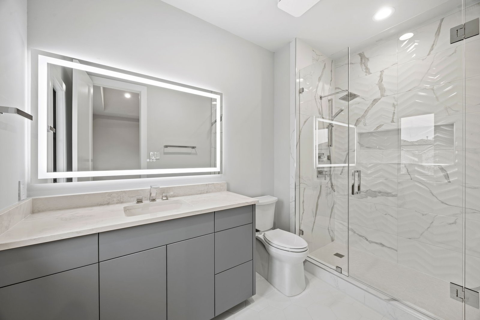 Renovated West Plano bathroom with a gray vanity, illuminated mirror and glass-enclosed shower